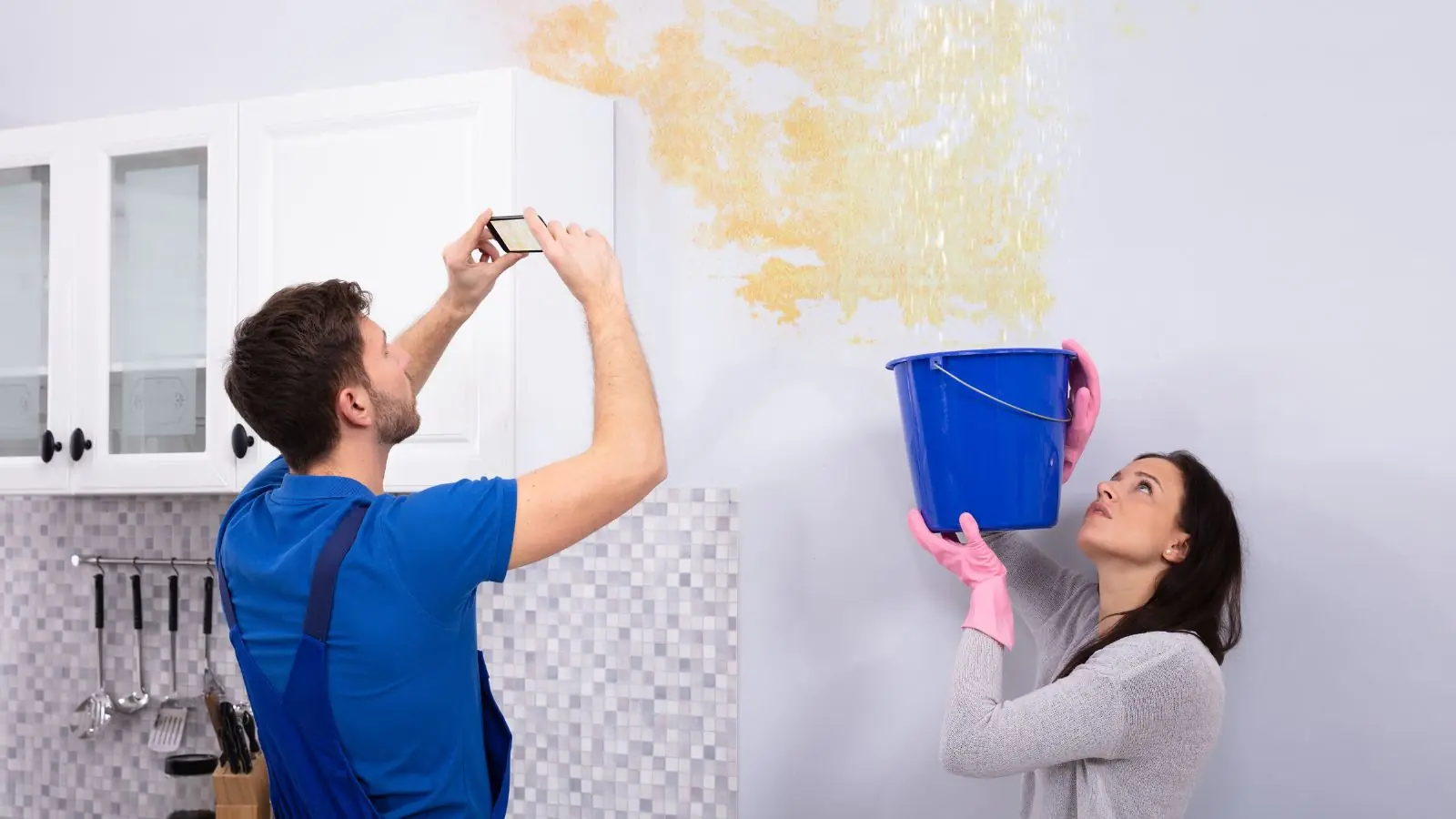 Homeowners checking a water-stained wall with a bucket and taking a photo of the damage
