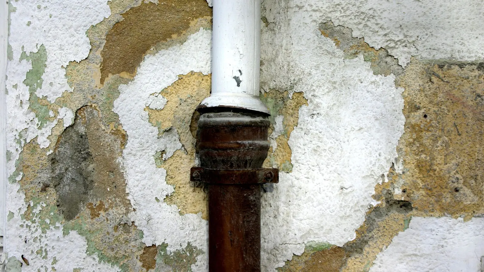 Wall with stained, flaking plaster around a rusted vertical pipe